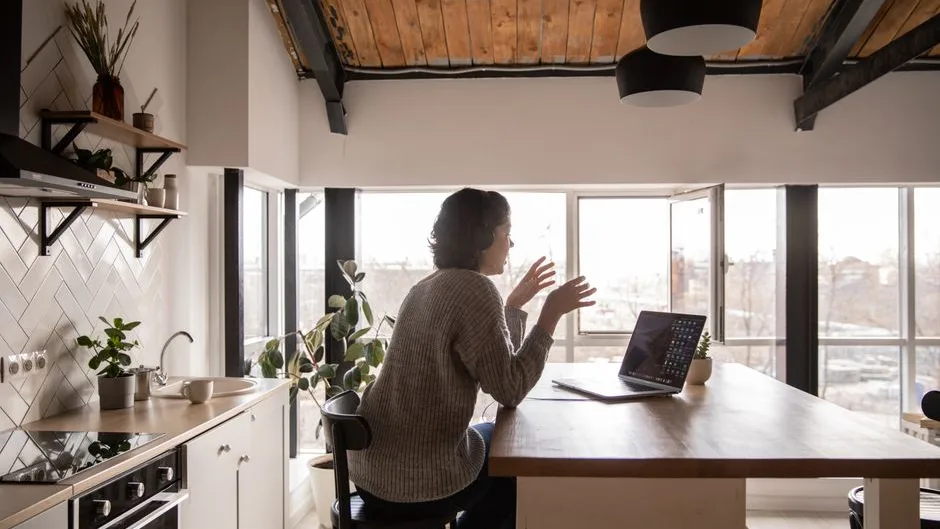 Femme vue de dos, assise à une table de cuisine lumineuse, qui échange en visio sur son ordinateur portable
